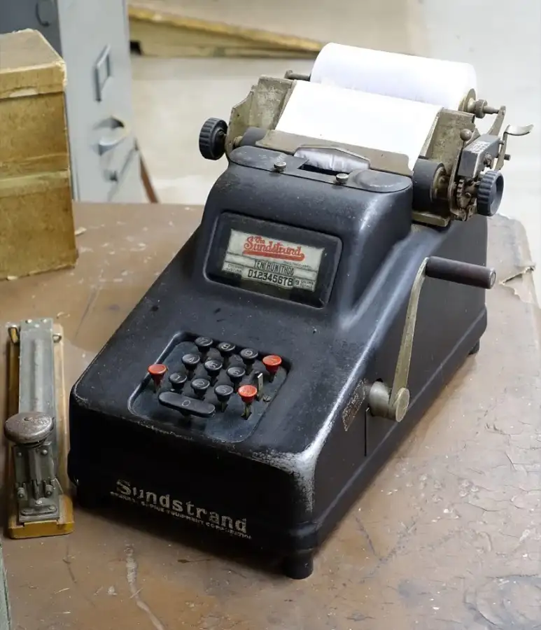 A black Sundstrand adding machine with a paper roll, on a wooden desk